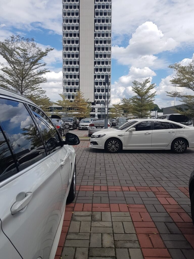 A car park filled with vehicles in the foreground, with a tall modernist tower block featuring a distinctive grid-patterned facade visible in the background, surrounded by trees under a partly cloudy sky — the building is the Malaysian Parliament complex in Kuala Lumpur.