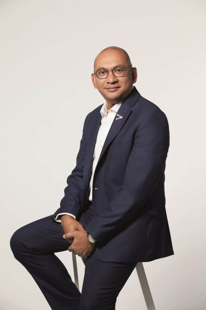 A bald man wearing glasses and a navy blue suit over a white shirt sits on a stool against a plain light grey background, with a small Accenture logo pin on his lapel